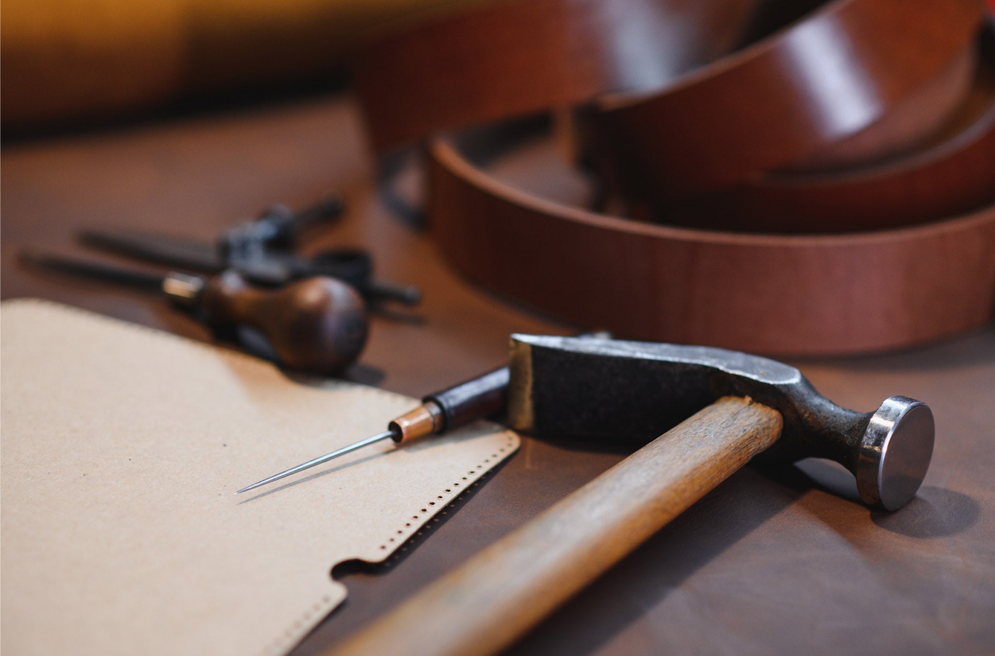 Leatherworking tools including a hammer, awl, and leather on a wooden surface.